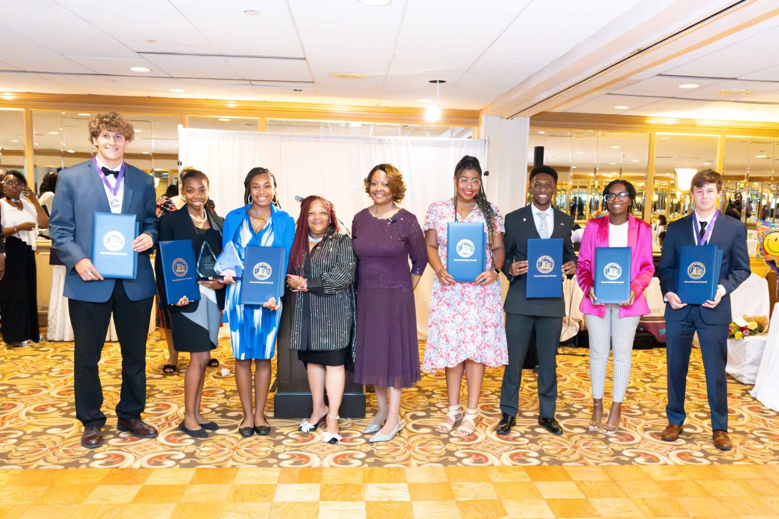 Group of award recipients and organizers standing in a row holding plaques indoors.
