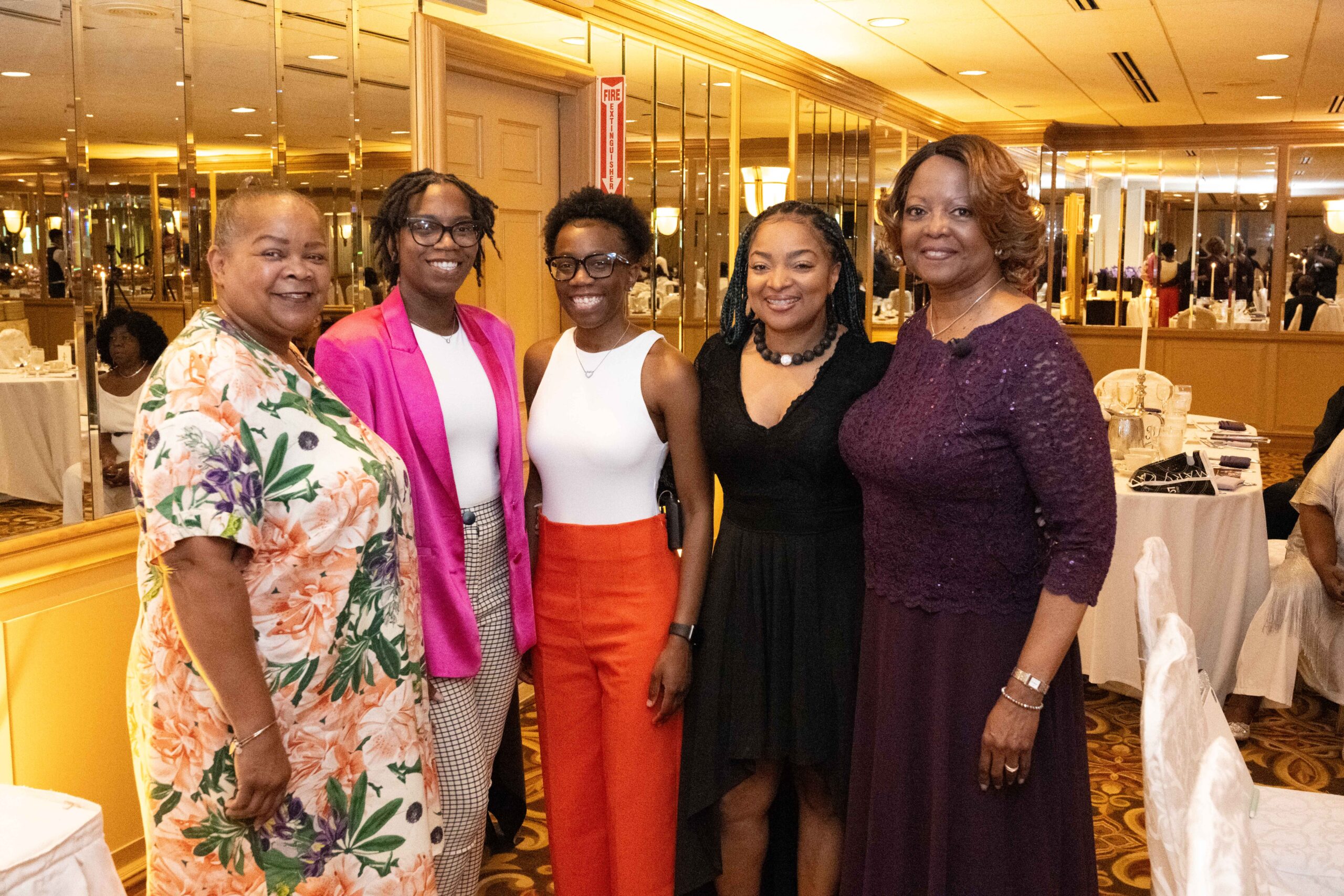 Five women dressed in colorful and elegant outfits posing indoors.